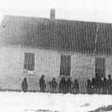 Black and white photo of a schoolhouse with children standing beside it