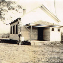 Photo of a white schoolhouse with a teacher