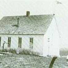 White schoolhouse with a flag, water pump, and chimney